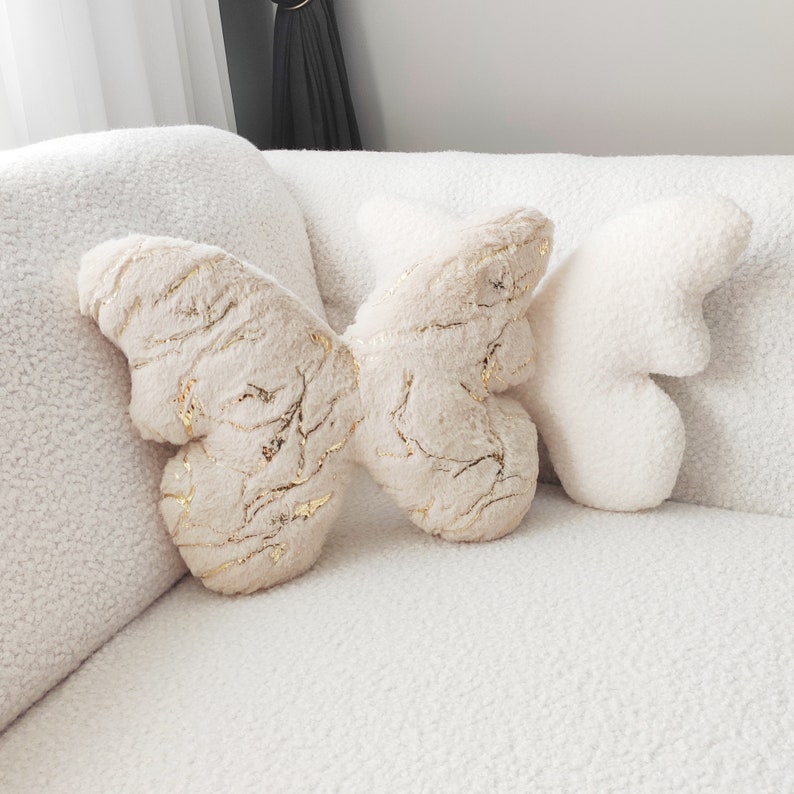 May include: Three butterfly-shaped decorative pillows in a neutral color palette. The central pillow has a beige, textured surface with gold vein-like accents. The other two pillows are white and fluffy, resting on a white, textured sofa.