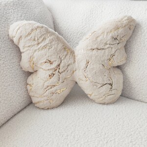 May include: A plush butterfly-shaped pillow in a cream color with gold metallic accents. The pillow is made of soft, textured fabric and is designed to resemble a butterfly. The pillow is resting on a white, textured surface.