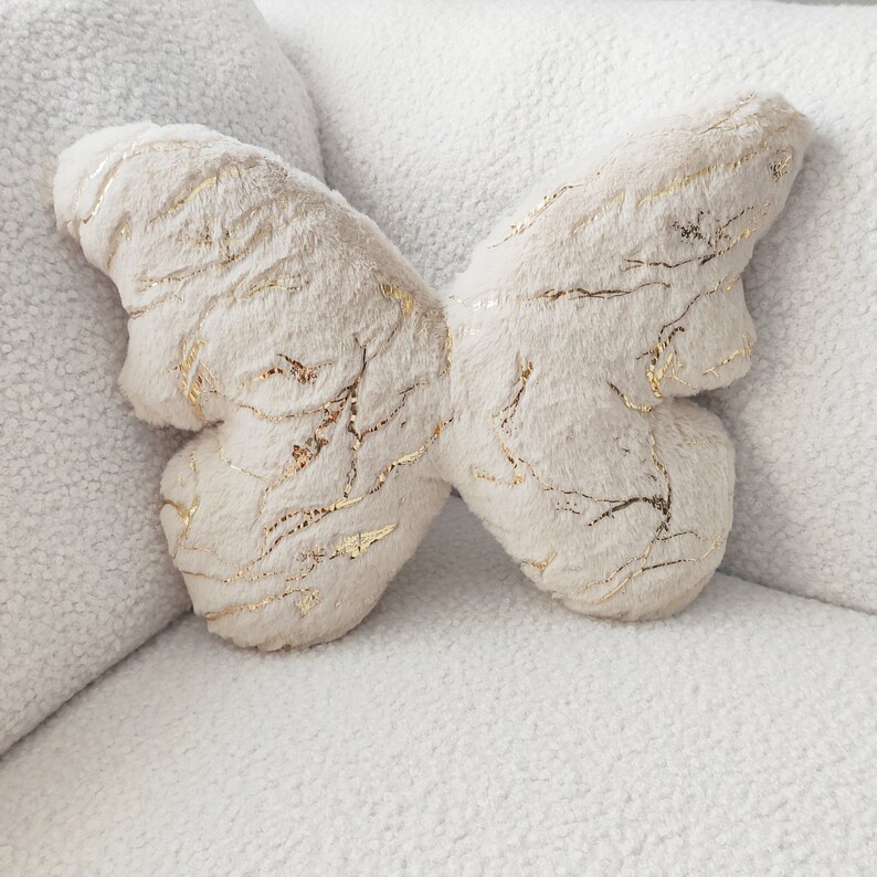 May include: A plush butterfly-shaped pillow in a cream color with gold metallic accents. The pillow is made of soft, textured fabric and is designed to resemble a butterfly. The pillow is resting on a white, textured surface.