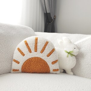 May include: A white, fluffy decorative pillow in the shape of a sun with orange and yellow accents. A white stuffed bunny with a green ribbon sits next to the pillow. The items are on a white, textured surface.