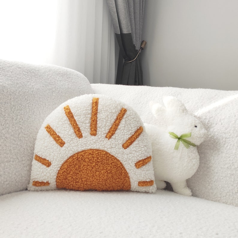 May include: A white, fluffy decorative pillow in the shape of a sun with orange and yellow accents. A white stuffed bunny with a green ribbon sits next to the pillow. The items are on a white, textured surface.