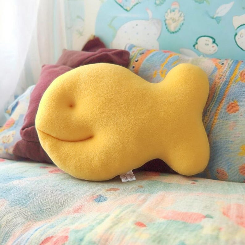 May include: A plush, yellow fish-shaped pillow with a smiling face rests on a bed. The pillow is the focal point, with a soft, textured appearance. Other pillows in shades of burgundy and blue with patterns are in the background.