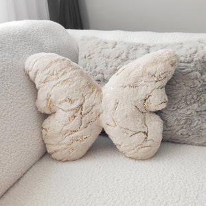 May include: A plush butterfly-shaped pillow in cream with gold metallic accents. The pillow is made of soft, textured fabric and is resting on a white couch with other decorative pillows.