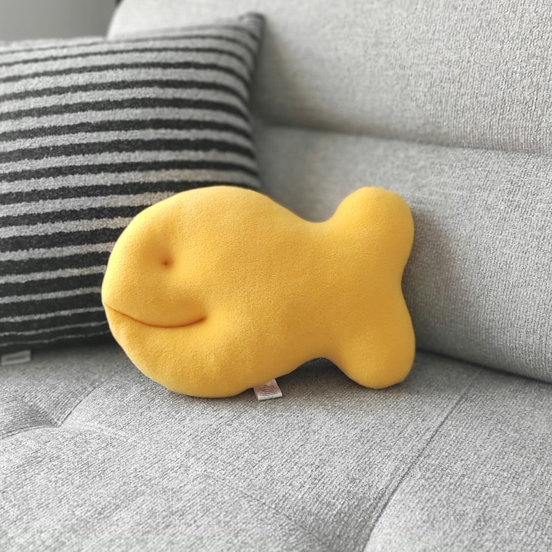 May include: A plush, yellow fish-shaped pillow with a smiling face rests on a gray sofa. A striped pillow is in the background. The fish pillow is a fun, whimsical decorative item.