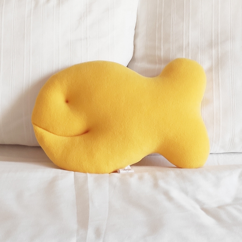 May include: A bright yellow, fish-shaped pillow rests on a white bed. The pillow has a simple, smiling face and a soft, plush texture. The pillow is a fun, decorative item for a bedroom or living space.