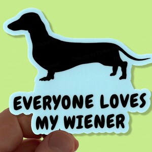 Könnte beinhalten: Aufkleber mit einer schwarzen Silhouette eines Dackelhundes und dem Text "EVERYONE LOVES MY WIENER" in Weiß auf hellblauem Hintergrund. Der Aufkleber wird von einer Hand gehalten.
