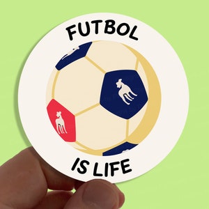 Pode incluir: Etiqueta redonda branca com uma bola de futebol branca e preta com uma silhueta de cão azul e branco em cada painel. O texto "FUTBOL IS LIFE" está impresso em preto na etiqueta.