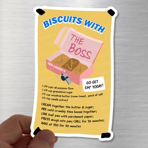 Peut inclure: Un autocollant illustré jaune et rose avec une recette de "Biscuits avec le Boss" inspirée de la série télévisée Ted Lasso. La recette comprend les ingrédients et les instructions pour faire des biscuits. L'autocollant présente une image de dessin animé d'une boîte rose avec deux biscuits à l'intérieur. Le texte "Go Get Em' Today!" est sur un rectangle blanc.