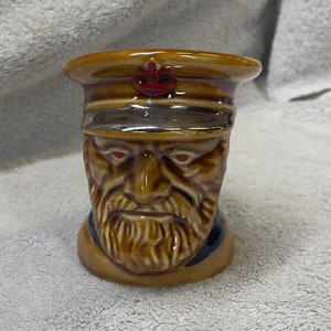 Puede incluir: Una taza de cerámica con forma de cabeza de hombre con barba marrón y sombrero rojo y plateado. La taza tiene ojos rojos y una expresión seria.