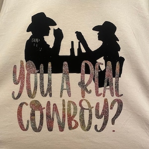 May include: A white t-shirt with a glittery design that says "You a real cowboy?" with a silhouette of two people in cowboy hats sitting at a bar.