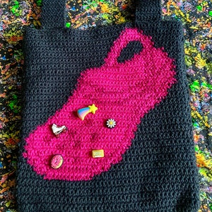 May include: Black crocheted tote bag with a large pink patch in the shape of a roller skate. The patch has several small colorful pins attached to it.