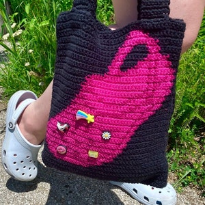 May include: A black and pink crocheted tote bag with a pink flamingo design. The bag has several colorful charms attached to it, including a rainbow, a daisy, a roller skate, and a donut.