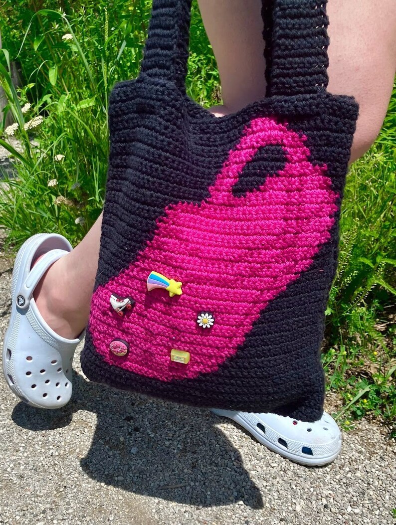 May include: A black and pink crocheted tote bag with a pink flamingo design. The bag has several colorful charms attached to it, including a rainbow, a daisy, a roller skate, and a donut.
