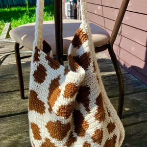 May include: A handmade crochet bag with a cow print design. The bag features a white base with brown spots, and a cream-colored strap. The bag is sitting on a wooden surface, with a chair and outdoor setting in the background.