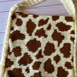 May include: Cream-colored crochet shoulder bag with a brown cow print pattern. The bag has a cream-colored strap and border. The bag is handmade and has a unique, rustic aesthetic.
