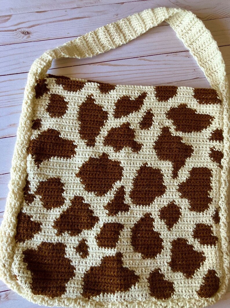 May include: Cream-colored crochet shoulder bag with a brown cow print pattern. The bag has a cream-colored strap and border. The bag is handmade and has a unique, rustic aesthetic.