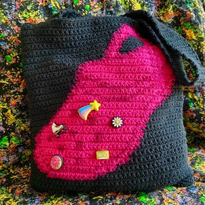 May include: A black and pink crocheted tote bag with a variety of colorful pins and patches attached to it. The bag has a large pink section with a black outline.