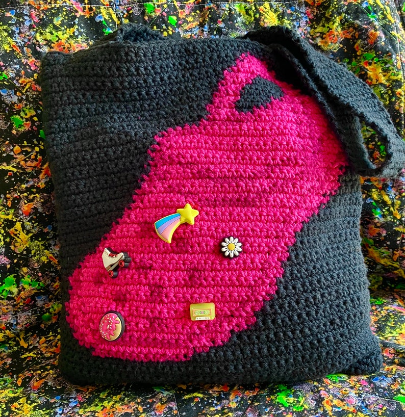 May include: A black and pink crocheted tote bag with a variety of colorful pins and patches attached to it. The bag has a large pink section with a black outline.