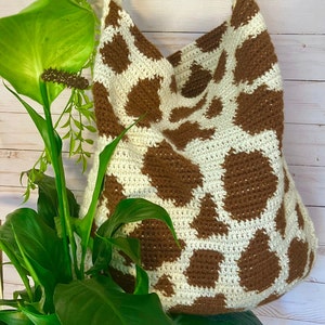 May include: A crochet tote bag featuring a cream-colored base with irregular brown patches in a cow-print pattern. The bag has a wide, integrated shoulder strap and a soft, textured construction made from yarn, displaying a consistent stitch.