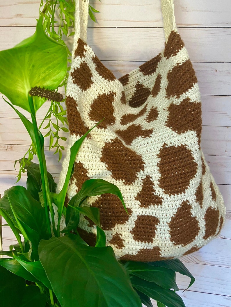 May include: A crochet tote bag featuring a cream-colored base with irregular brown patches in a cow-print pattern. The bag has a wide, integrated shoulder strap and a soft, textured construction made from yarn, displaying a consistent stitch.