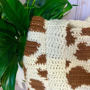 May include: Crocheted bag with a cow print pattern. The bag features a cream-colored base with brown spots. The crochet stitches are visible, adding texture to the design. The bag is partially visible, with a green plant in the background.