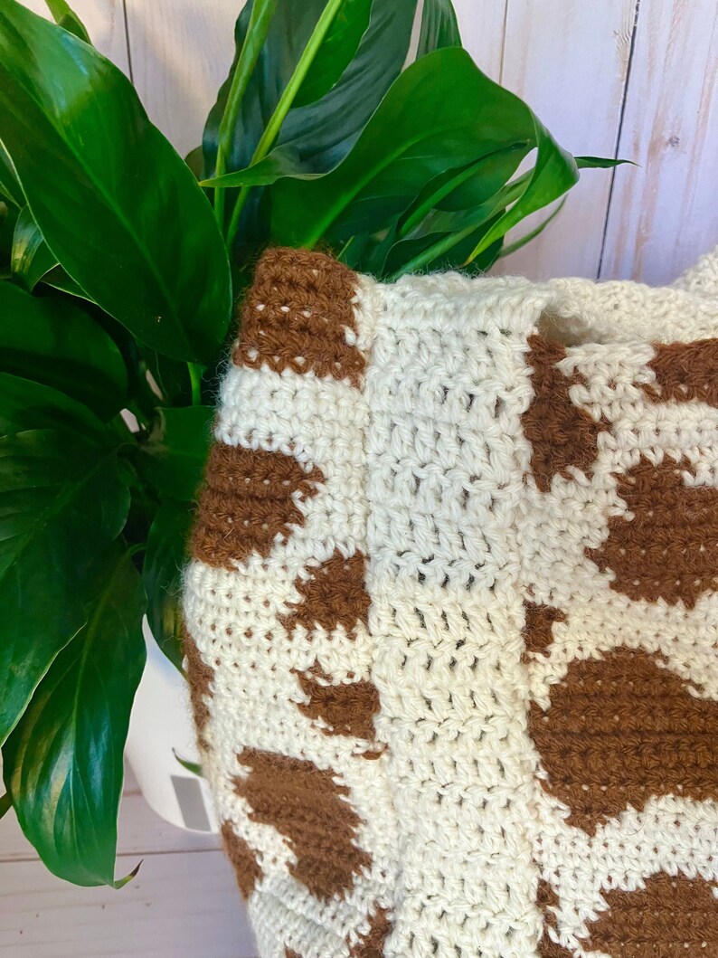 May include: Crocheted bag with a cow print pattern. The bag features a cream-colored base with brown spots. The crochet stitches are visible, adding texture to the design. The bag is partially visible, with a green plant in the background.