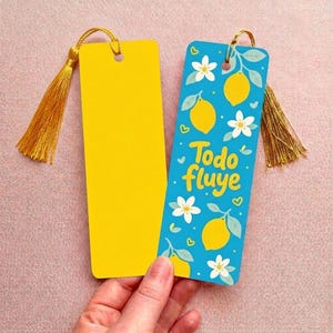 Puede incluir: Dos marcapáginas rectangulares con borlas doradas. Uno es de color amarillo sólido. El otro es azul con un patrón de limones amarillos, flores blancas y pequeños corazones verdes, con el texto amarillo Todo fluye en el centro.