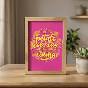 Op de afbeelding: Een ingelijste print met een felroze achtergrond en gouden letters. De tekst luidt "Cada pétalo que coloreas es un paso hacia la calma", wat zich vertaalt naar "Elk bloemblad dat je kleurt is een stap naar rust". De lijst is gemaakt van lichtgekleurd hout.