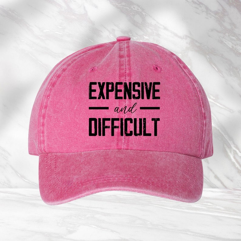 May include: Pink cotton baseball cap with the text "EXPENSIVE and DIFFICULT" in black.