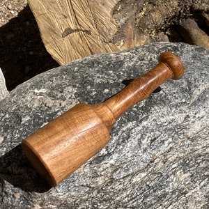 May include: A wooden mortar and pestle. The pestle has a rounded handle and a cylindrical body. The mortar is a wide, cylindrical shape. The wood has a warm, natural tone, with visible grain. A kitchen utensil.