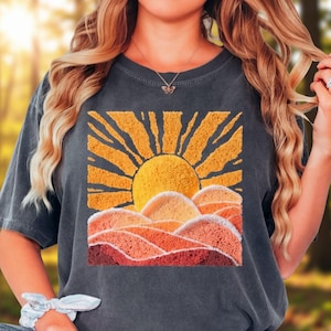 May include: Dark gray t-shirt featuring a vibrant embroidered design of a sun rising over layered hills. The sun is a bright yellow, with orange and red hues in the hills. The shirt is a relaxed fit.
