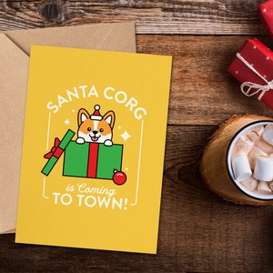 May include: A yellow greeting card with the text "SANTA CORG is Coming TO TOWN!" features a cartoon corgi wearing a Santa hat. The card is next to a brown envelope, a mug of hot chocolate with marshmallows, and wrapped gifts.