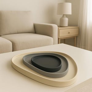 May include: Three nested, organic-shaped serving trays in a neutral color palette. The largest tray is a soft cream, the middle one is gray, and the smallest is dark charcoal. The trays are arranged on a white table, with a sofa and lamp in the background.