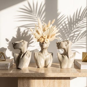 May include: Three beige ceramic head and shoulder sculptures arranged in a row. The center sculpture holds a bouquet of dried beige grasses, palm leaves, and small flowers. The left sculpture features a hand resting on the chest area.