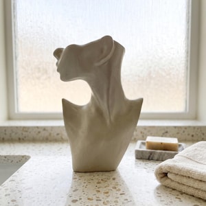 May include: A beige ceramic bust sculpture depicting the head and shoulders of a human figure in profile. The sculpture features a smooth, matte finish with a hollow top opening, showcasing a neutral tone and a stylized, abstract anatomical form.