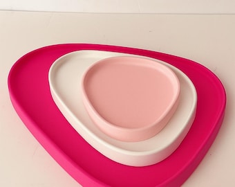 May include: A nested set of three ceramic trays in shades of pink and white. The largest tray is a vibrant fuchsia, followed by a white tray, and a smaller, pale pink tray. The trays have a smooth, matte finish and a modern, organic shape.