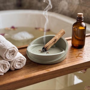 May include: A light sage green concrete incense holder containing a burning palo santo stick with visible smoke, positioned next to three rolled white cotton towels and a small amber glass bottle with a cork stopper on a wooden tray.