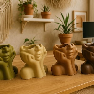 May include: Four ceramic face planters in shades of green, beige, tan, and brown sit on a wooden table. The planters have a unique design, with a face shape and hands supporting the chin. Plants and decor are in the background.