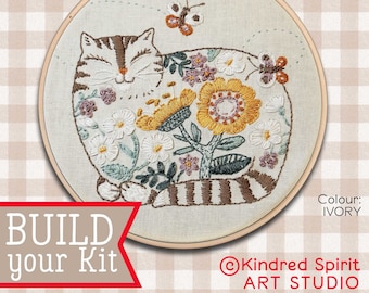 Pode incluir: Um aro de bordado bege com um desenho de um gato dormindo rodeado de flores. O gato é marrom e branco com listras. As flores são amarelas, brancas e rosas. O aro tem um diâmetro de 22 cm. O texto "BUILD your Kit" está no canto inferior esquerdo. O texto "Kindred Spirit ART STUDIO" está no canto inferior direito. O texto "Colour: IVORY" está no canto inferior direito.