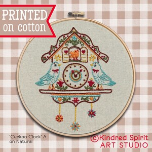 May include: A printed embroidery pattern of a cuckoo clock with two birds on either side. The clock is decorated with flowers and has a yellow, red, and blue color scheme. The pattern is printed on natural cotton fabric and is ready to be stitched.