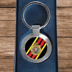 May include: Silver-colored keychain with a round emblem featuring a crest design with red, yellow, and black stripes. The keychain is presented in a dark blue box, set against a wooden background.