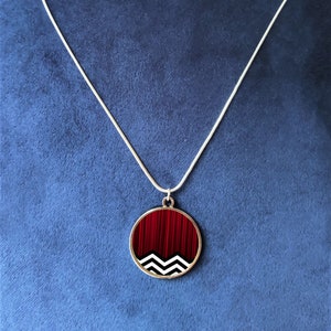Puede incluir: Un collar de cadena de plata con un colgante redondo que presenta un patrón geométrico rojo y blanco. El colgante tiene un diseño en zigzag blanco y negro sobre un fondo rojo.