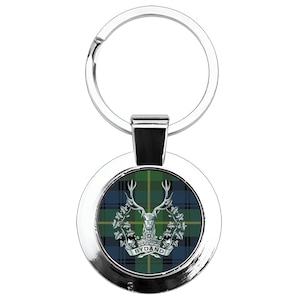 May include: A silver-coloured keyring with a circular pendant featuring a green, blue, and black tartan pattern. The centre displays a stag emblem and the word "BYDAND". The keyring has a silver ring for keys.