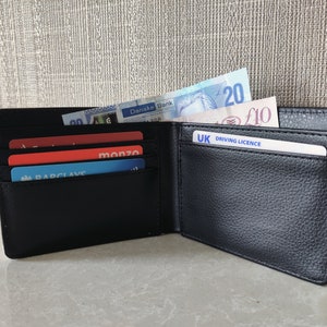 May include: A black leather wallet with multiple card slots and a billfold. The wallet is open and contains several credit cards, a driver's license, and some cash. The cash includes a twenty euro bill and a ten pound bill.