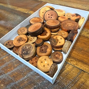 May include: A white tray filled with various sizes of wooden buttons. The buttons are natural wood tones, ranging from light tan to dark brown, with visible grain and drilled holes for sewing. Some buttons are stacked.
