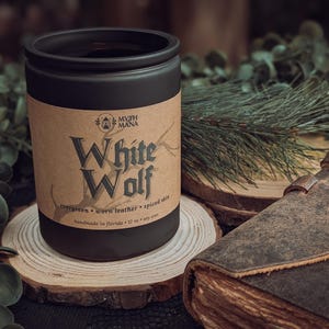 May include: A black candle jar with a label that reads "White Wolf" in a gothic font. The label also lists the scent notes: evergreen, worn leather, and spiced skin. The candle sits on a wooden slice, with a leather-bound book and greenery in the background.