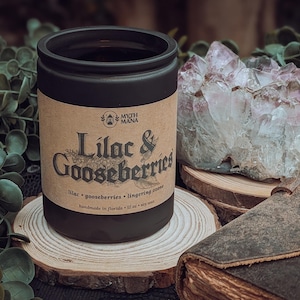 May include: A black candle jar with a label that reads "Lilac & Gooseberries" in a vintage font. The candle sits on a wooden slice with a crystal cluster and a leather-bound book in the background. The candle is 12 oz.