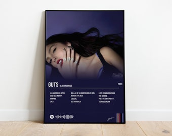 May include: A poster for Olivia Rodrigo's album "GUTS" featuring a close-up of her face. The poster is dark blue with white text and a barcode. The album title "GUTS" is in large white letters at the top of the poster. The album's tracklist is listed below the title.