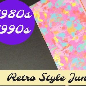 May include: A pink and orange handmade journal with a marbled cover featuring pink, blue, and yellow swirls. The cover also has the text "Handmade Journal" and "1980s 1990s" printed on it. The journal is labeled as a "Retro Style Junk Journal".
