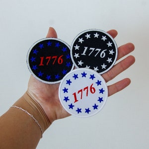 May include: Three round embroidered patches with a circle of blue stars around the number 1776 in red. The patches are black, white, and white with a blue border.